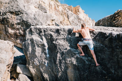 Full length of shirtless man climbing on rock against sky