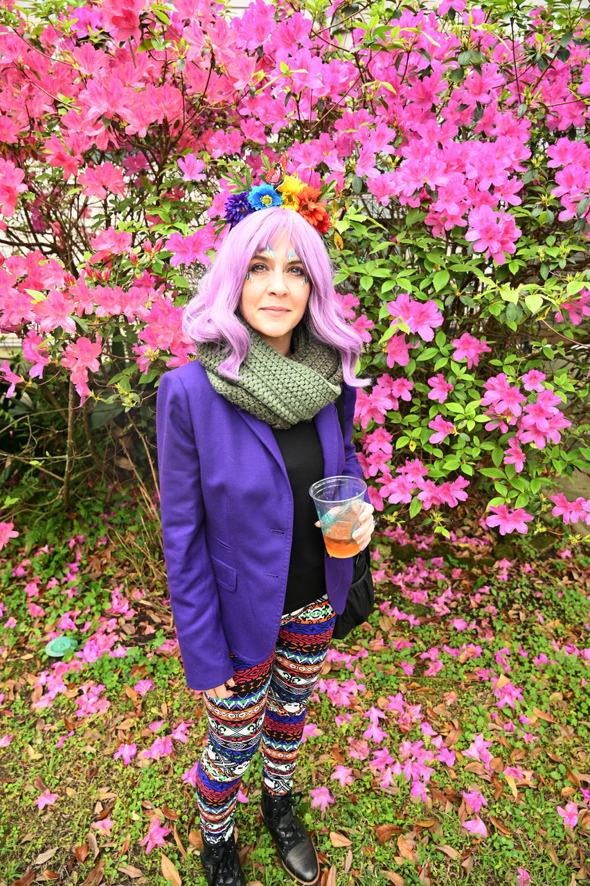 flowering plant, flower, one person, plant, pink color, young adult, front view, young women, standing, leisure activity, real people, lifestyles, beauty in nature, nature, looking at camera, day, clothing, women, casual clothing, beautiful woman, outdoors, warm clothing, scarf, flower head