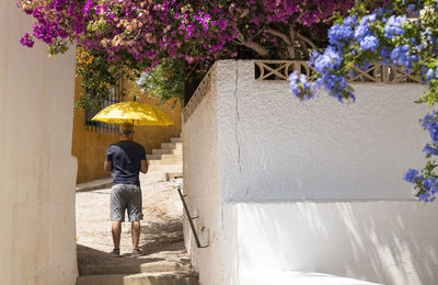 Rear view of adult man with yellow umbrella standing on street by white wall with flowers plants