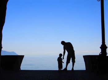 Silhouette father and son standing on beach against clear sky