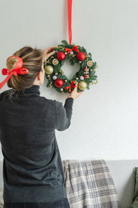 Woman hangs a coniferous wreath on the wall above the sofa, preparing for christmas at home.