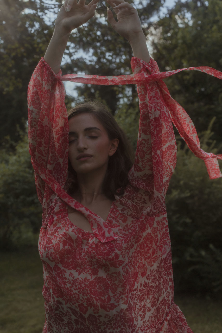 one person, red, tree, front view, women, young adult, adult, waist up, nature, portrait, clothing, standing, arm, leisure activity, plant, day, brown hair, arms raised, forest, hairstyle, dress, pink, outdoors, lifestyles, long hair, focus on foreground, spring, looking, limb, three quarter length, land, casual clothing