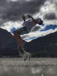 Full length of young man doing backflip over sand against cloudy sky