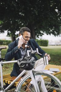 Happy businessman talking on mobile phone while sitting by bicycle at park