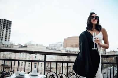 Low angle view of woman wearing sunglasses and champagne flute standing by railing against sky