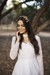 Charming tender young brunette in stylish white lace wedding dress and bridal wreath touching cheek and smiling while standing near tree in park