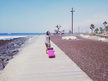 Full length of woman pulling luggage on footpath against sea during sunny day