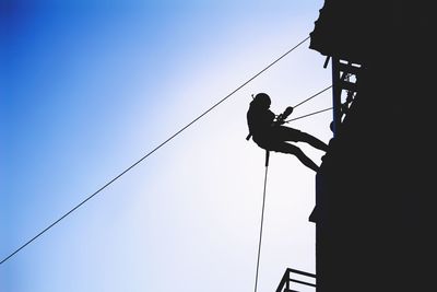 Low angle view of silhouette teenage boy climbing on building against sky during sunny day