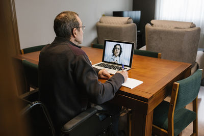 Senior man writing prescription sitting on wheelchair consulted by female doctor through video call at home