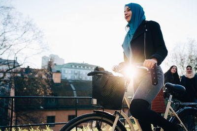 Back lit of young woman cycling by female friends against sky in city