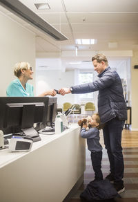 Side view of mature man giving id card to doctor while son standing by reception in hospital