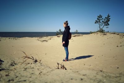 Side view of young woman standing on sand against blue sky