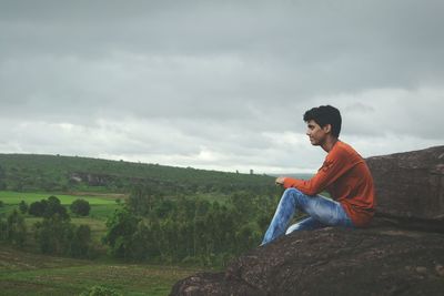 Side view of man sitting on rock against cloudy sky