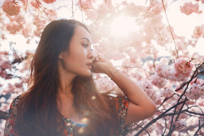 Young asian woman, in a floral pattern dress, in front of blossoming cherry tree in the spring time