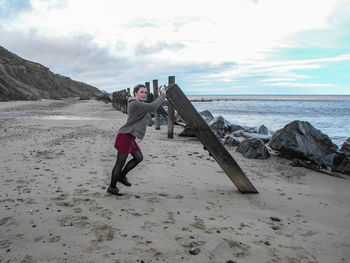 Portrait of young woman standing while holding wooden post at beach