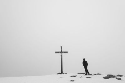 Man standing on snow covered hill by religious cross against sky during winter