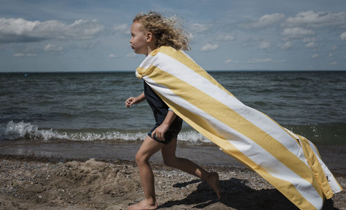 Side view of girl wearing cape while running at beach against sky during sunny day