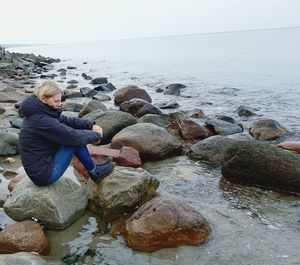 Boy sitting on pebbles at beach against clear sky