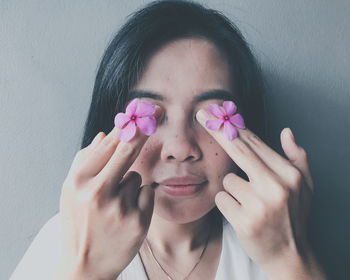 Close-up of woman covering eyes with pink periwinkle flowers against wall