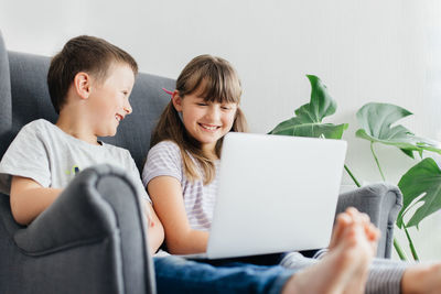 A schoolgirl with her brother are watching studying on a laptop while sitting in an armchair. 