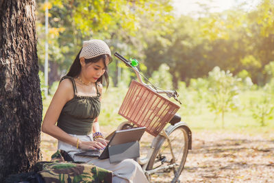 Asian woman in green singlet working with laptop under the tree next to a white bicycle in a park