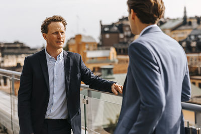 Mature male entrepreneur talking with colleague while standing near railing in balcony