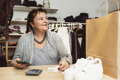 Smiling cashier counting with smart phone on checkout table in clothing store