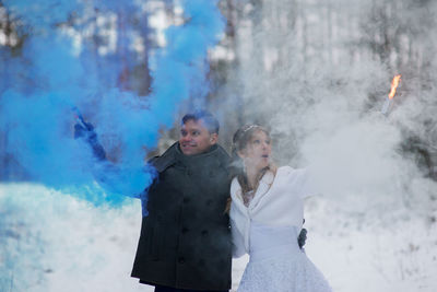 Smiling wedding couple holding distress flares while standing at forest during winter