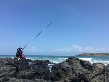 Fishing rod on rock by sea against blue sky