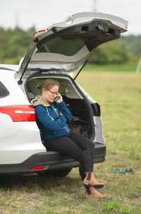 Woman talking on phone while sitting in car trunk on field