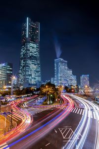 Light trails on road by illuminated buildings against sky at night