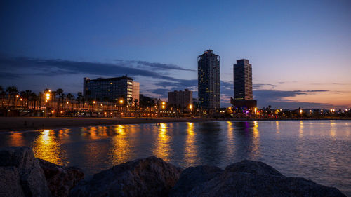 River by illuminated buildings against sky at dusk