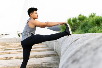 Side view of fit asian male athlete warming up and stretching legs before workout in city while standing near stone fence and looking away