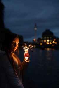 Portrait of young woman holding illuminated string light against sky at night