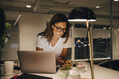 Confident female entrepreneur using smart phone while sitting with laptop at illuminated desk in workplace during night