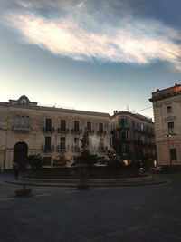 Fountain by street against buildings in city at sunset