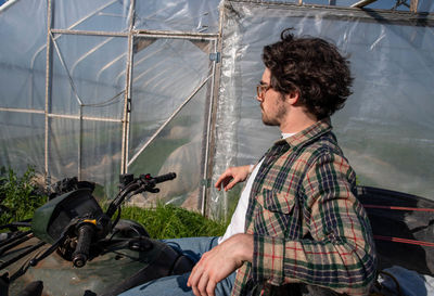 Young farmer in flannel shirt sits on off road vehicle outside greenhouse