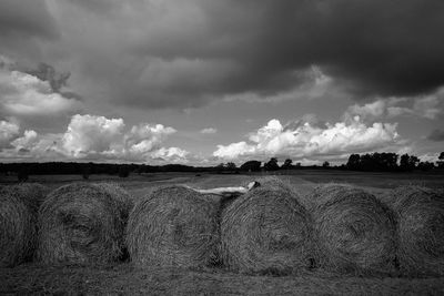 Side view of naked woman lying on hay bales at farm against cloudy sky