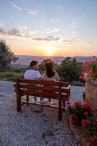 Rear view of couple sitting on bench at sunset