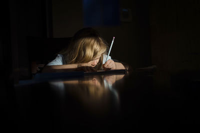 Girl writing homework while sitting at table in darkroom in house