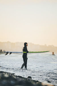 Surfer with surfboard standing in sea at sunset