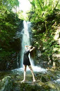 Full length of young woman standing by waterfall in forest