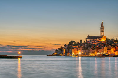 Illuminated buildings by sea against sky during sunset