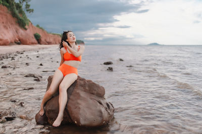Young woman drinking water while sitting on rock at beach