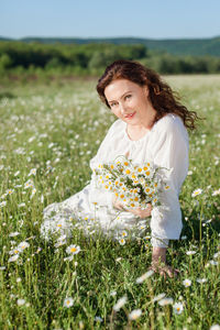 A laughing, relaxed woman in a summer dress sits among a blooming field of daisies on summer day