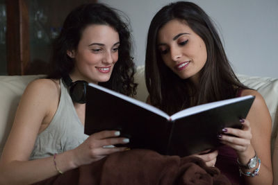 Young female friends reading book while sitting on couch at home