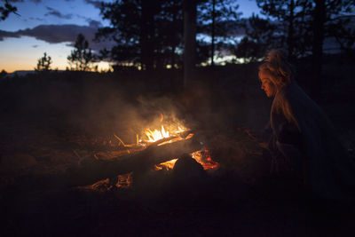 Woman sitting by bonfire in forest at night