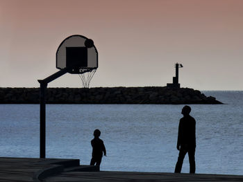 Silhouette men standing by basketball hoop against sky during sunset