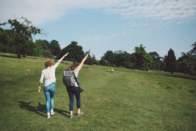Rear view of friends with arms raised standing on grassy field against sky
