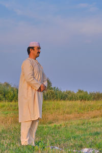 A muslim senior man wearing a skullcap and traditional white clothes prays in an agricultural field